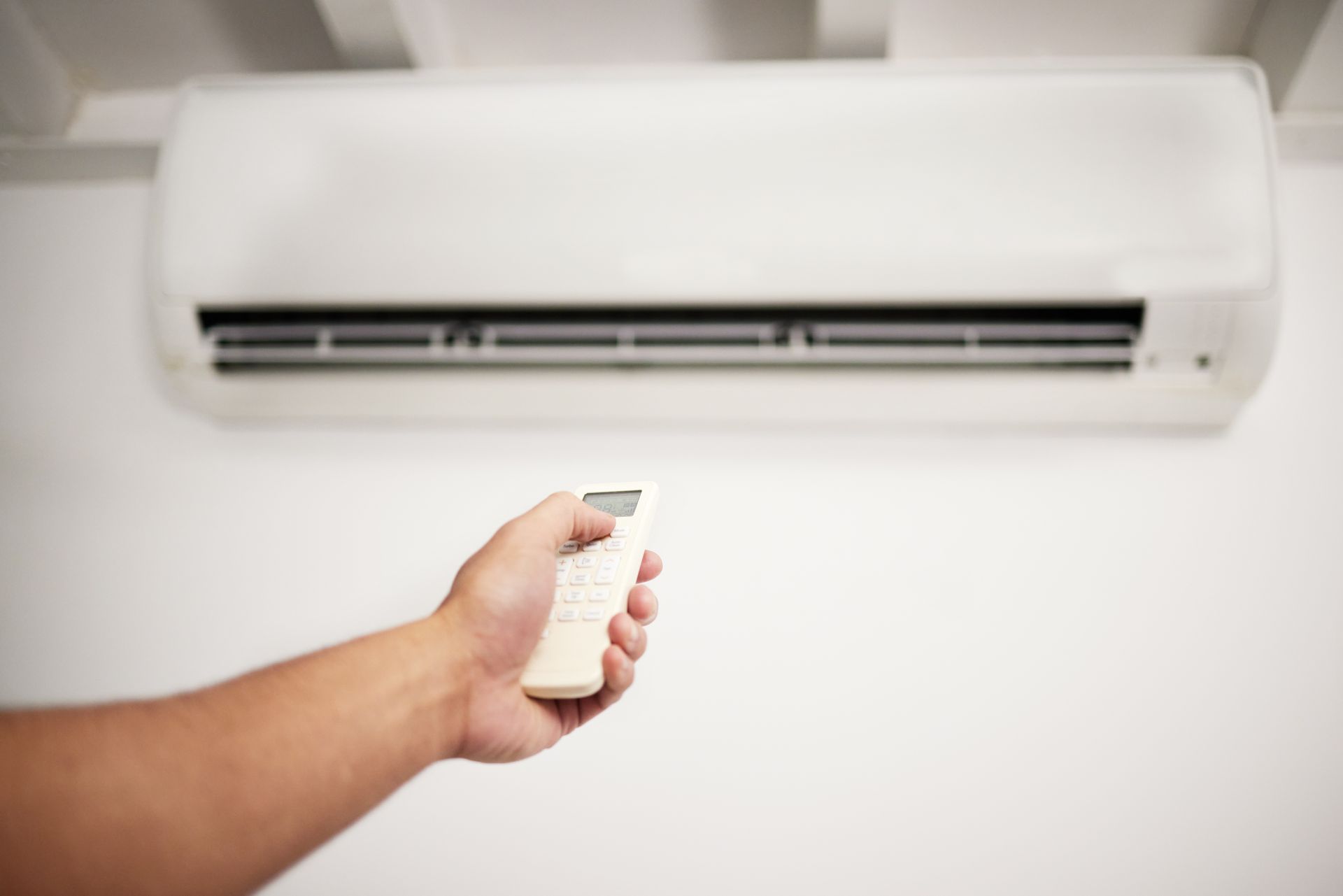 A hand holding a white remote control pointed at a white wall-mounted air conditioner unit A hand holding a white remote control pointed at a white wall-mounted air conditioner unit