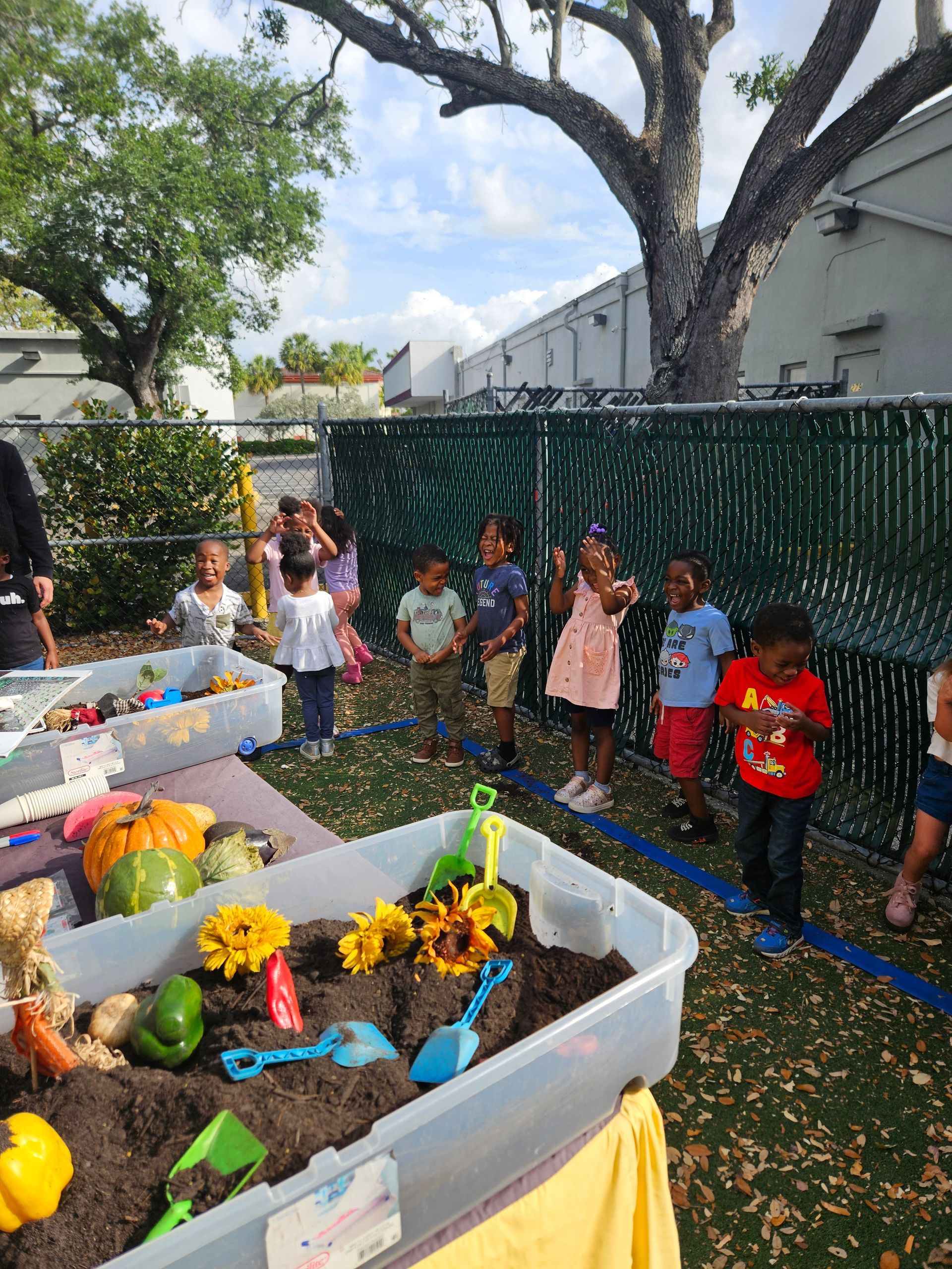 Children playing outdoors at a garden-themed sensory table with sunflowers, soil, and toys.