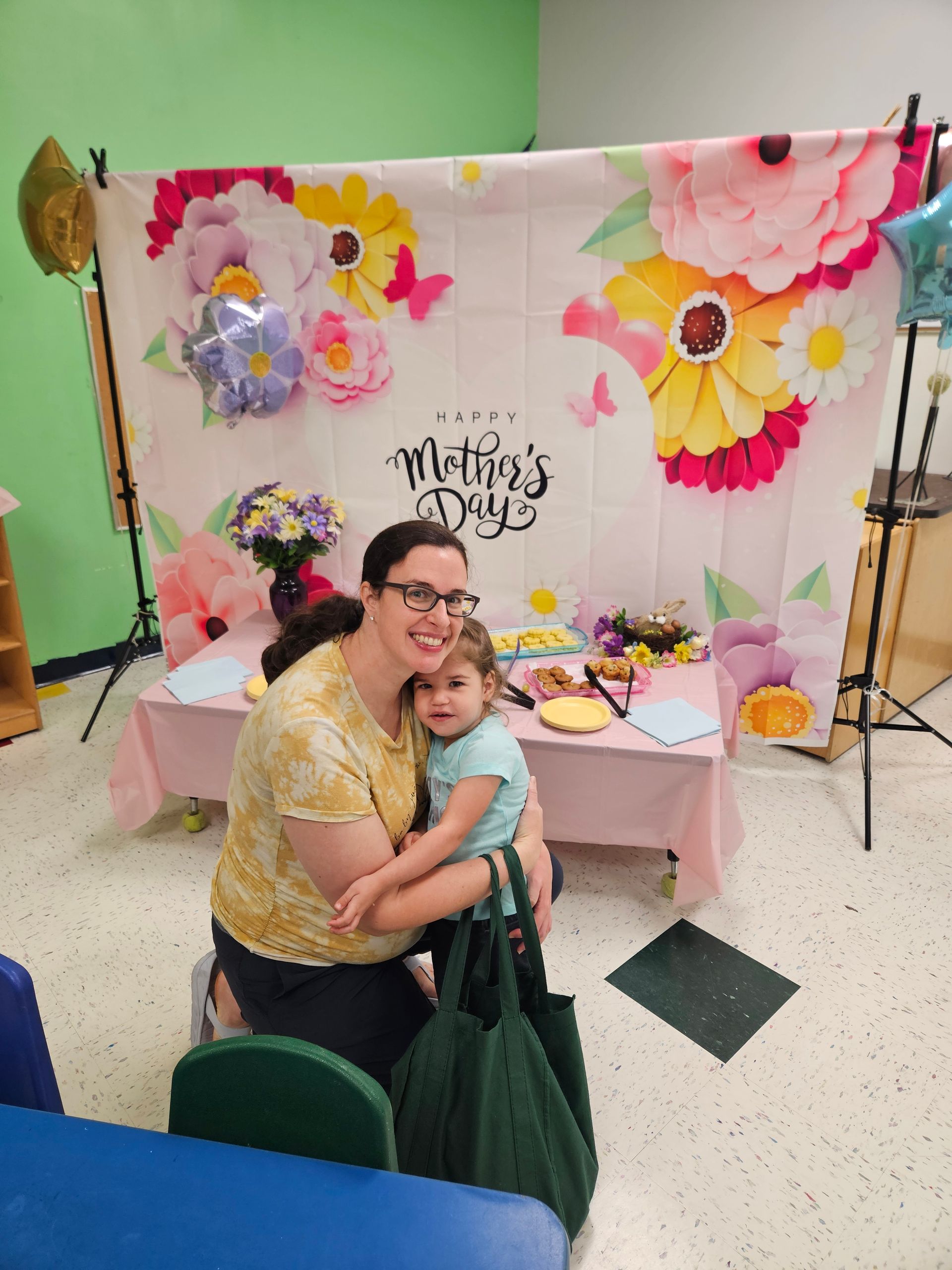 Woman embraces a child at a Mother's Day celebration. Floral backdrop, table with treats.