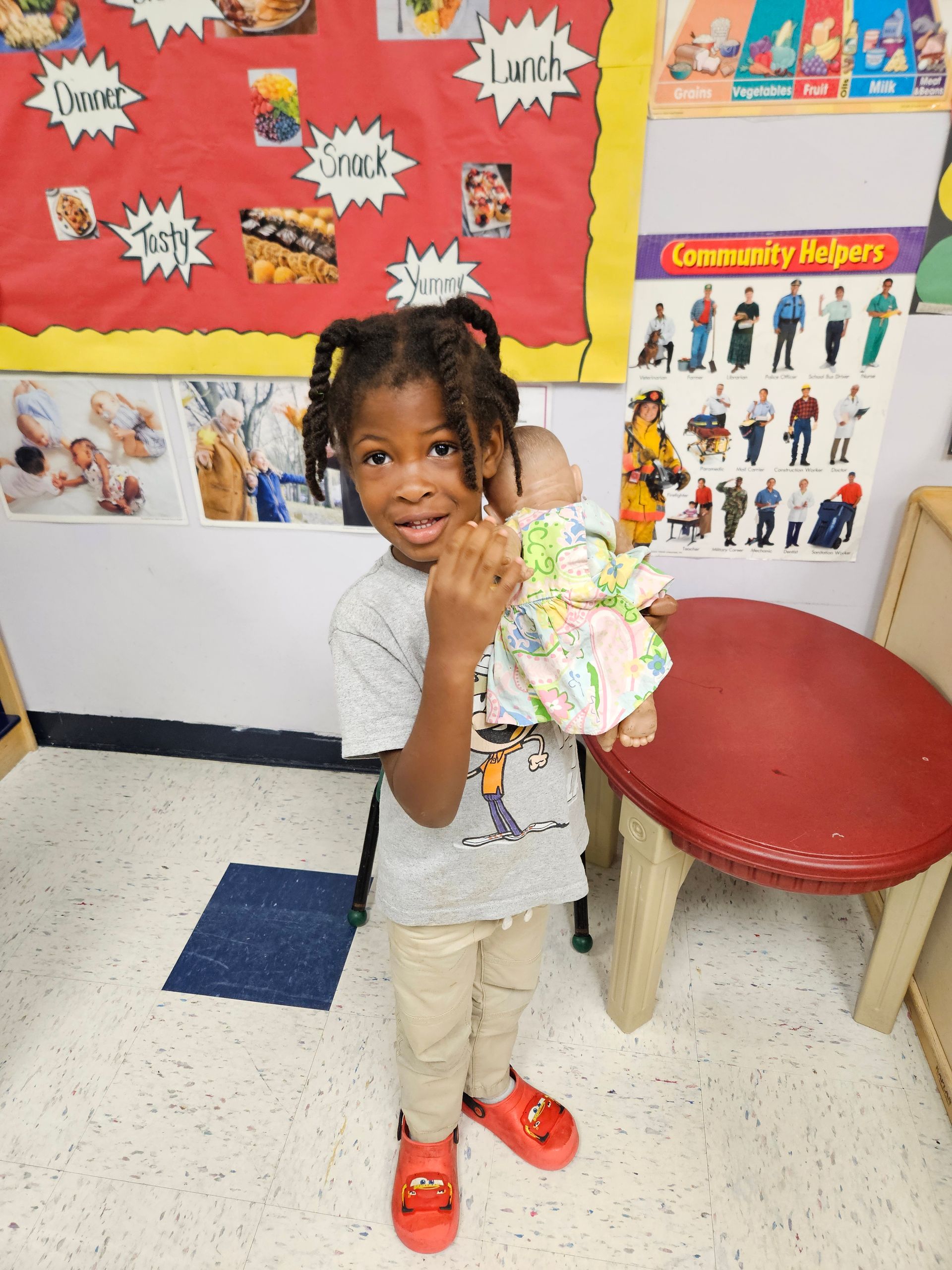 Young child with pigtails holds a doll, smiles in a classroom with a bulletin board and table.
