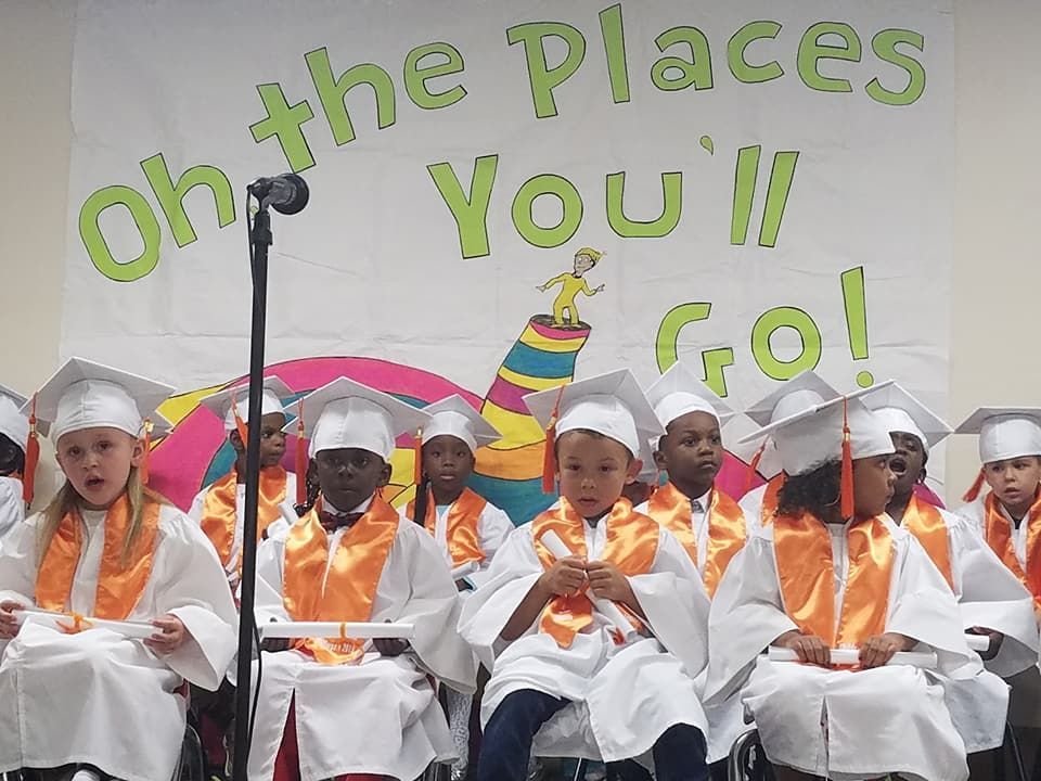 Children in graduation robes and caps, performing on stage, banner says 
