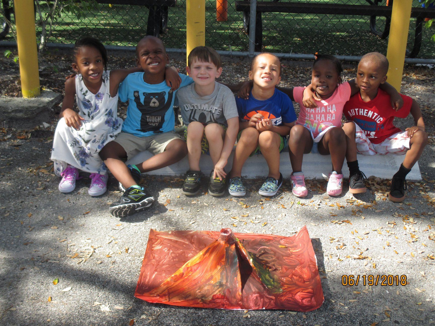 Six children smiling, sitting on a ledge; holding an art project of a volcano in a park.