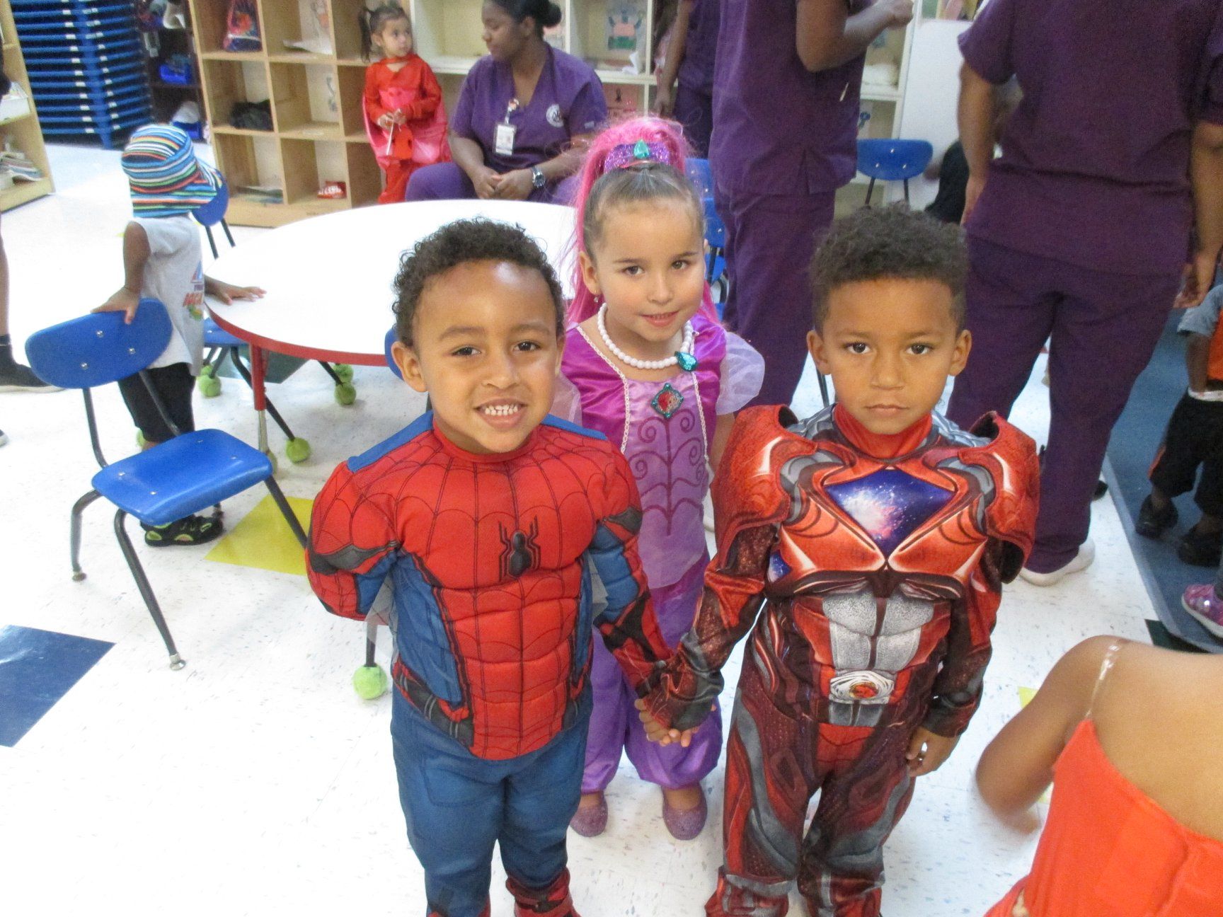 Three children in costumes smiling at the camera: Spider-Man, a princess, and a superhero. Classroom setting.