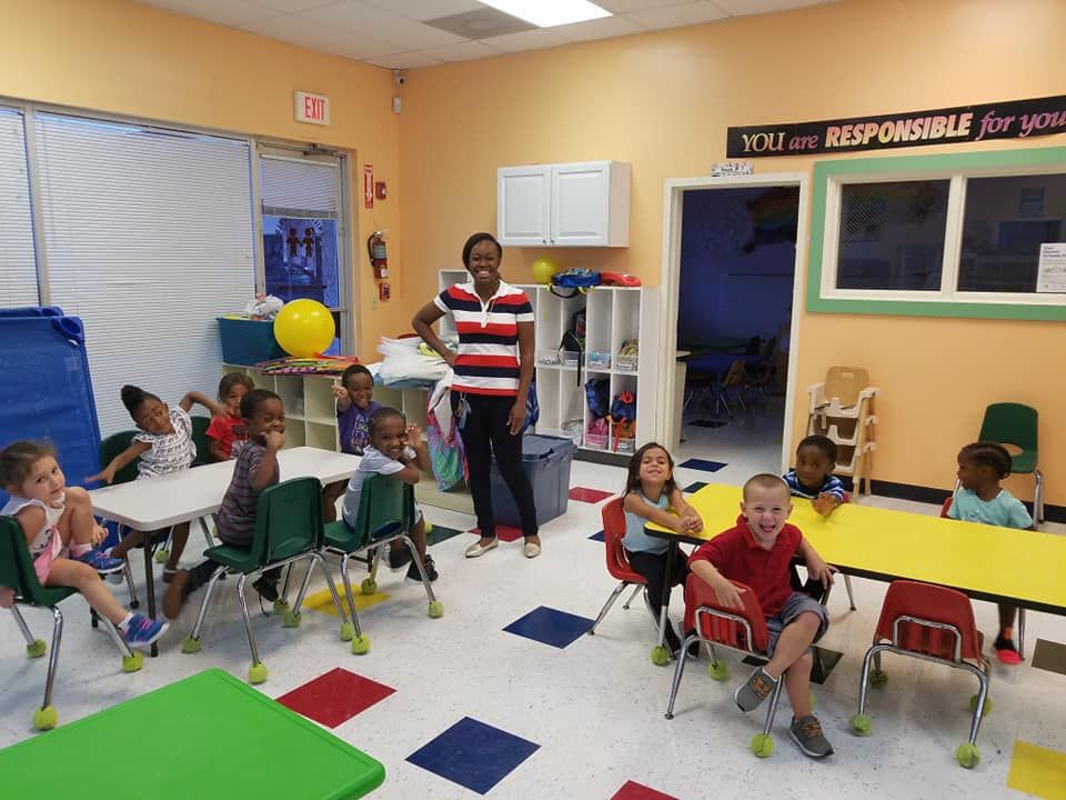 Children and teacher in a colorful classroom. Children seated at tables, teacher standing.