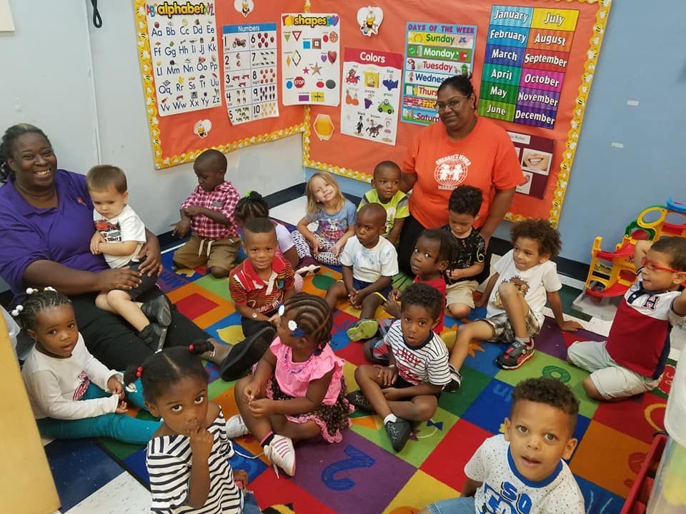 Children and two teachers in a brightly colored classroom. Smiling kids sit on a multi-colored rug.