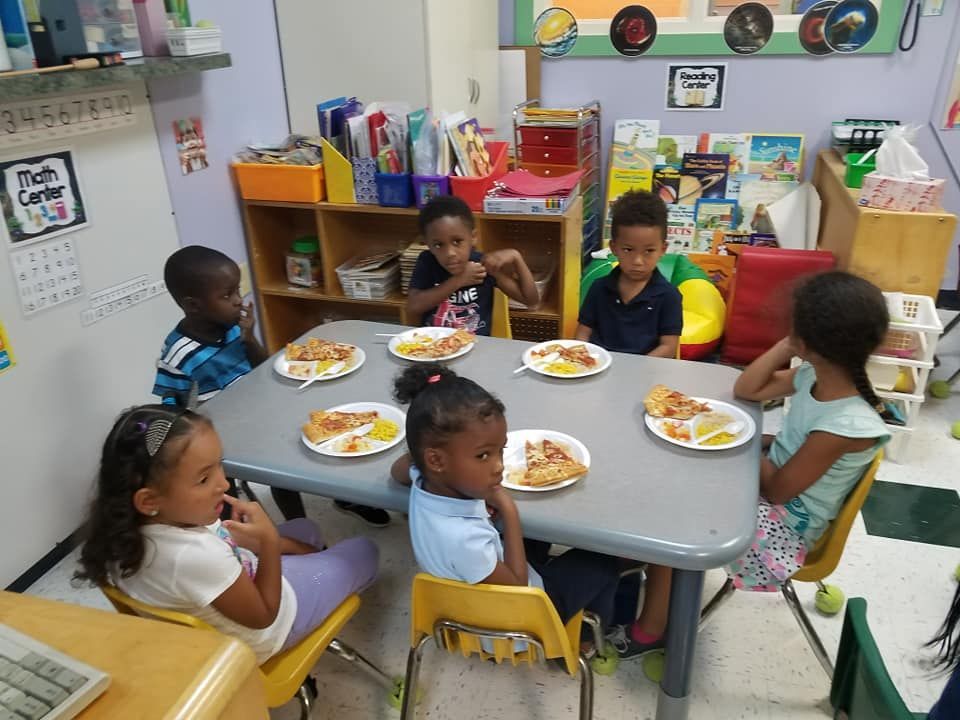 Children seated at a table eating pizza in a colorful classroom setting.