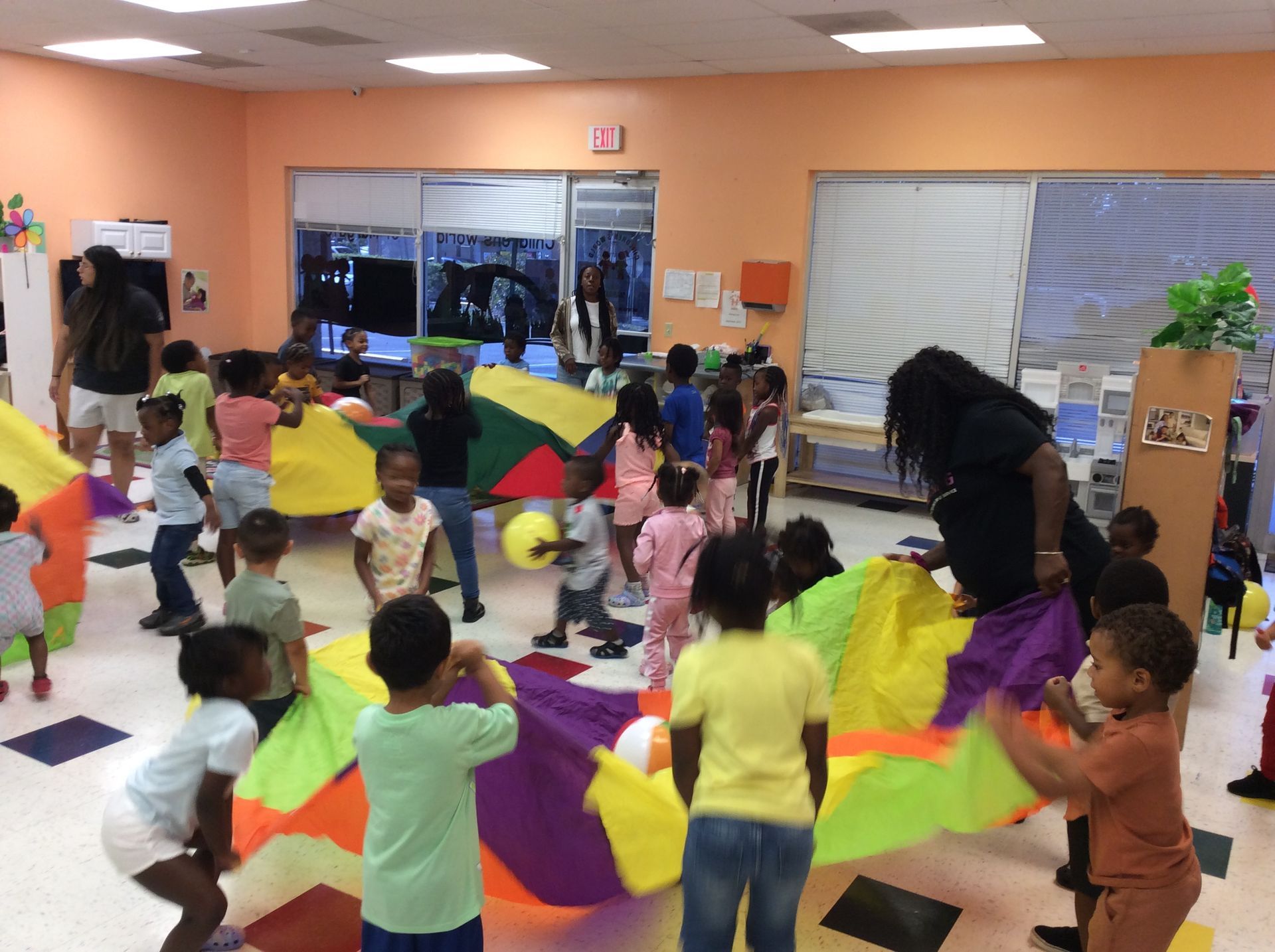Children playing with a colorful parachute in a brightly lit room with adult supervision.
