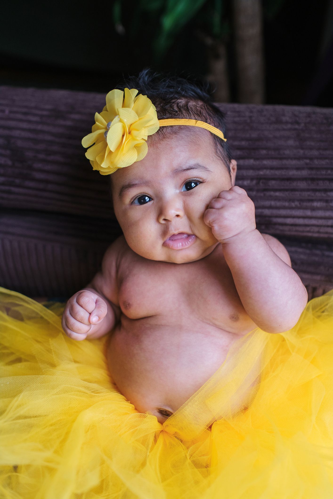 Baby in a yellow tutu and matching headband sitting on a couch, touching their face.