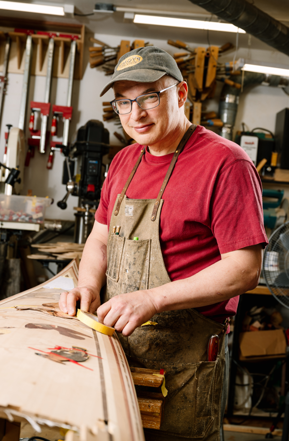 Person in a workshop wearing a red shirt and cap, working on a wooden board at a workbench.