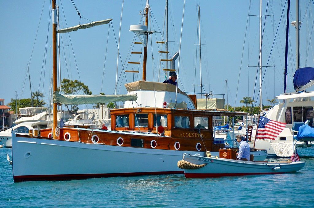 Wooden sailboat docked at a marina with blue water and other yachts in the background.