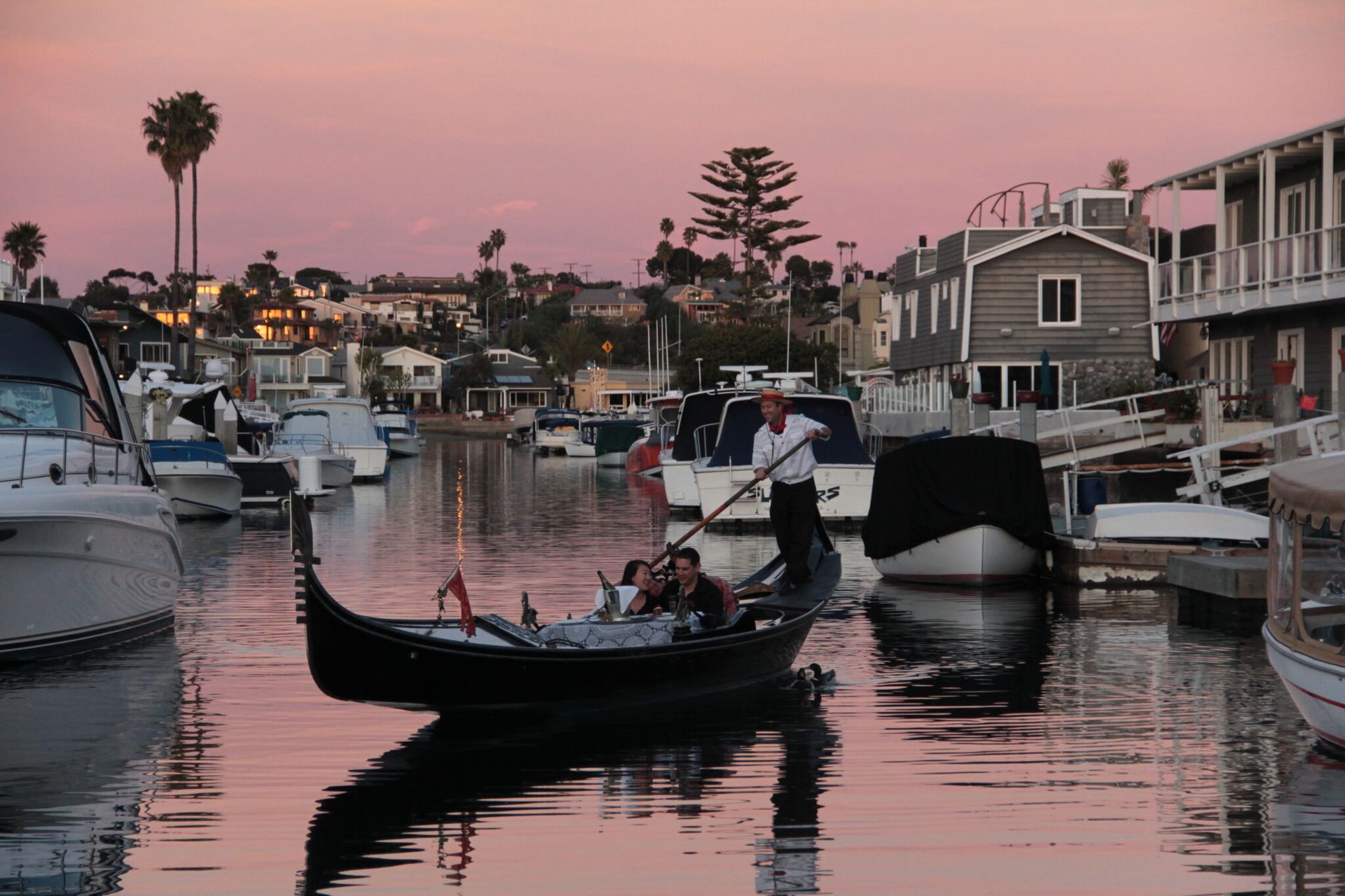 Gondola on a calm harbor at sunset, with boats and waterfront houses reflecting in pink water.