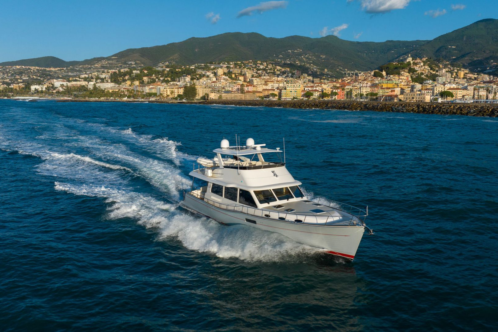 White motor yacht speeding across blue water with a coastal city and hills in the background.