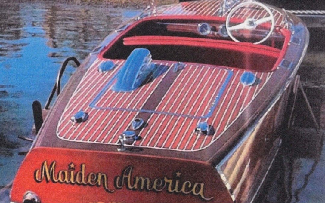 Red speedboat with wooden deck and blue seats, named “Maiden America,” floating on calm water