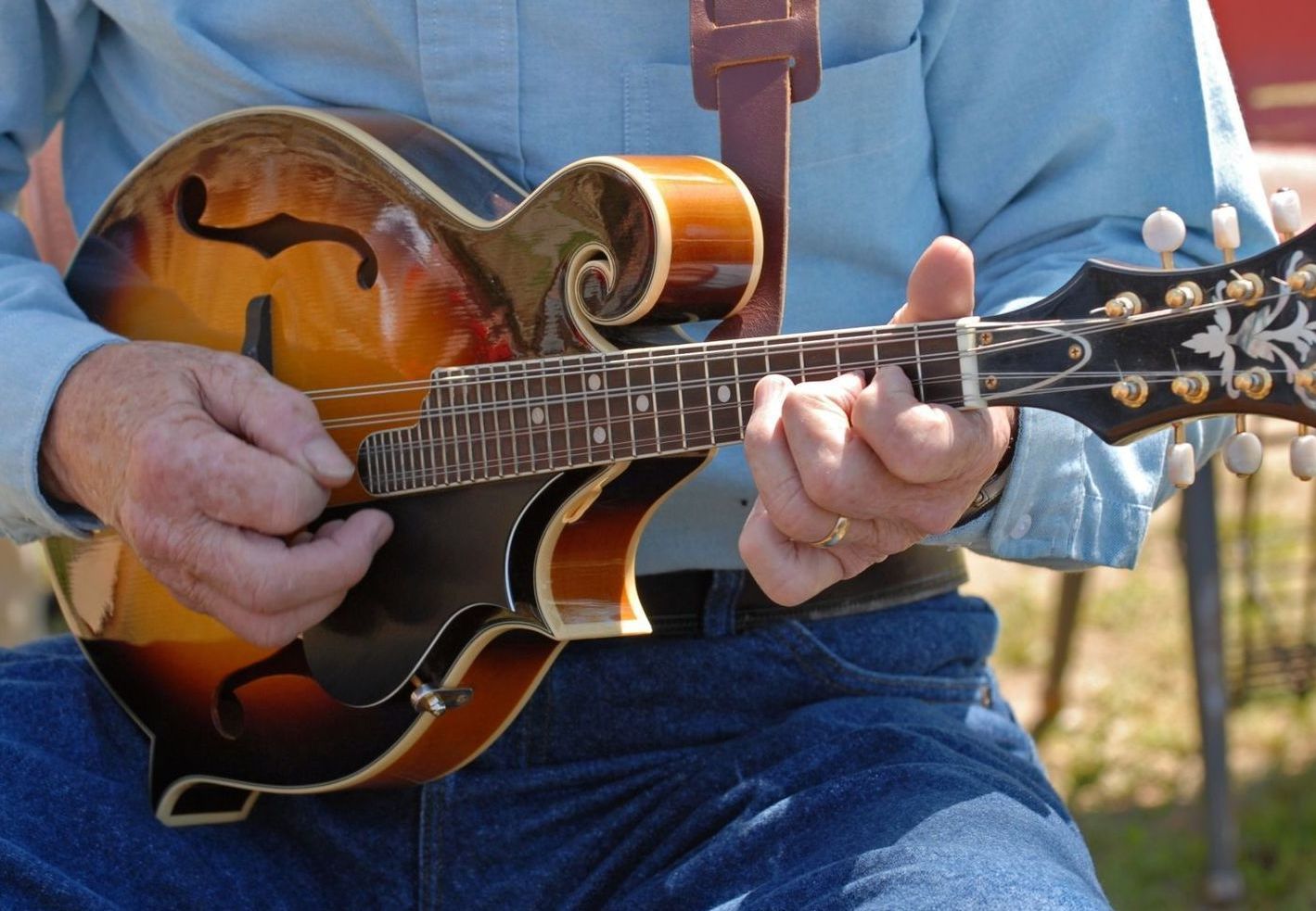 Person playing a sunburst mandolin outdoors while seated in jeans and a blue shirt