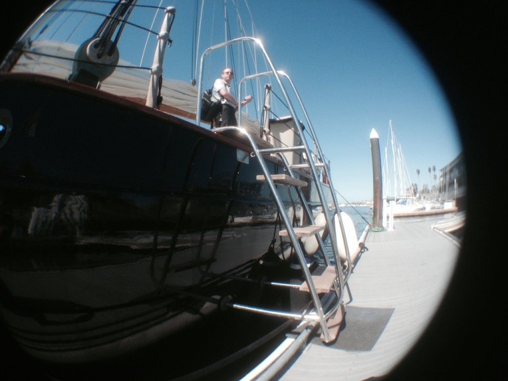 Person on ladder beside a black sailboat docked at a marina with city skyline in the background