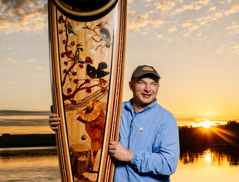 Male artist smiling beside a tall decorated canoe at sunset by a lake.