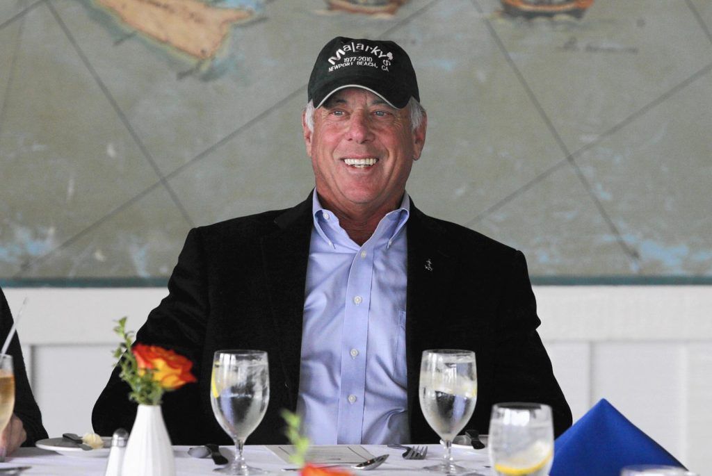 Smiling man in a black cap and jacket seated at a table with glasses and a small flower arrangement
