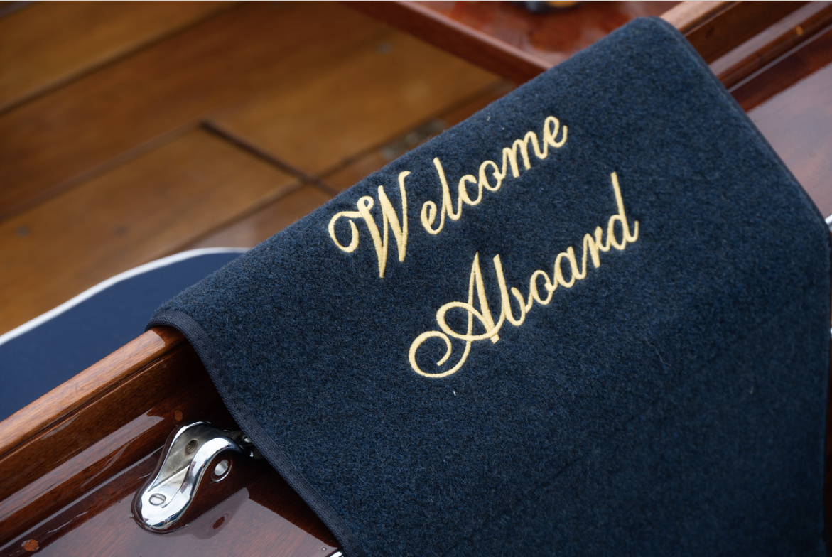 Navy towel embroidered “Welcome Aboard” draped over a polished wooden rail aboard a boat