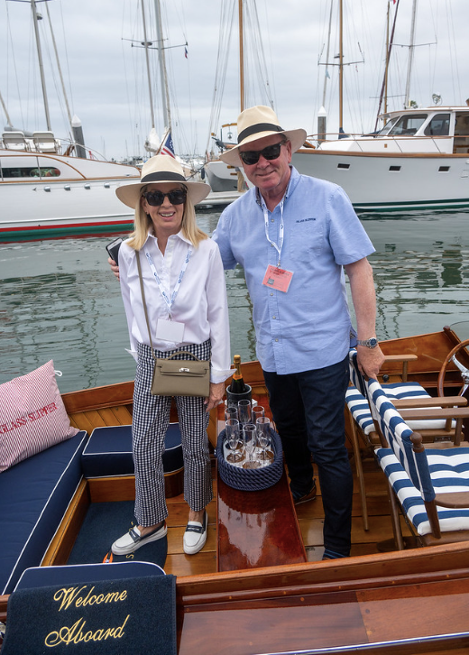 Two people posing on a boat deck by a marina, with sailboats in the background.