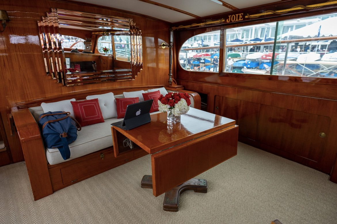 Wood-paneled boat cabin with white sofa, wooden table, and harbor view through windows.
