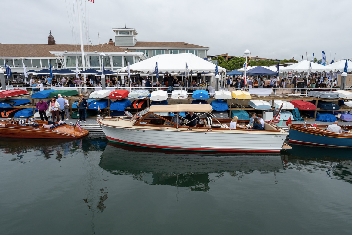Row of colorful fishing boats docked on calm water at a busy harbor with tents and buildings in the background