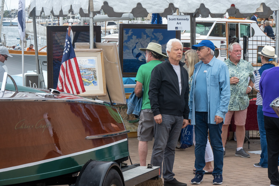 People gathered at a boat show beside a wooden boat with an American flag on a rainy dock.
