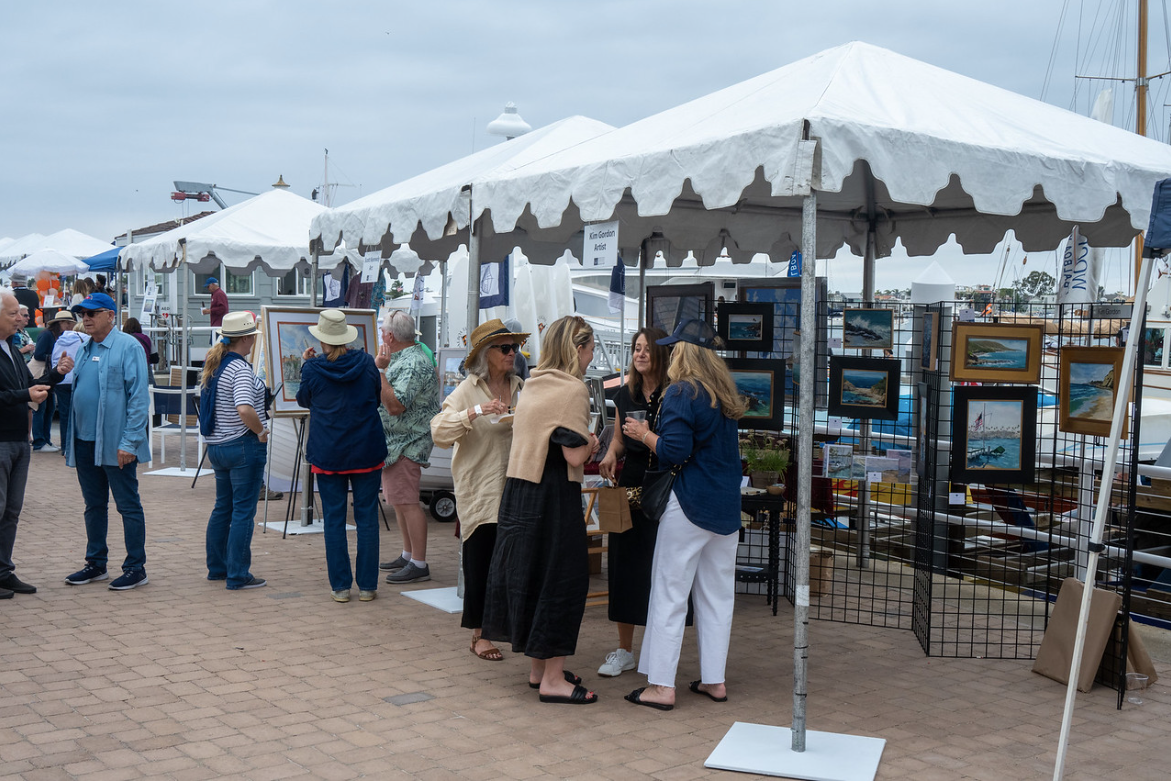 Outdoor art fair with tents, framed paintings, and people browsing displays along a brick walkway.