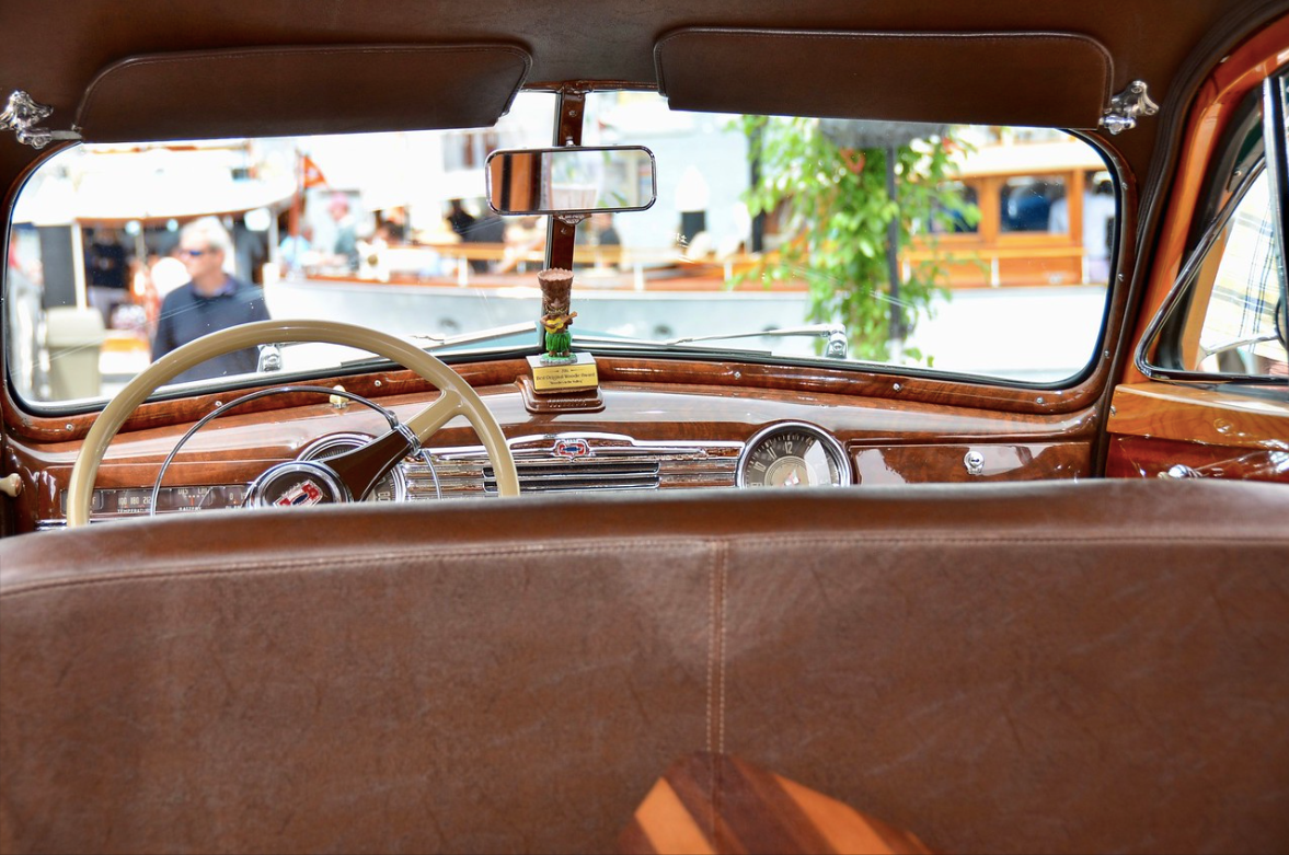 Interior of a vintage car with brown leather seats, dashboard, and rearview mirror, seen from the back seat.