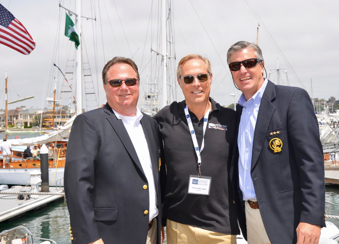 Three men in blazers posing at a marina, with boats and an American flag in the background.