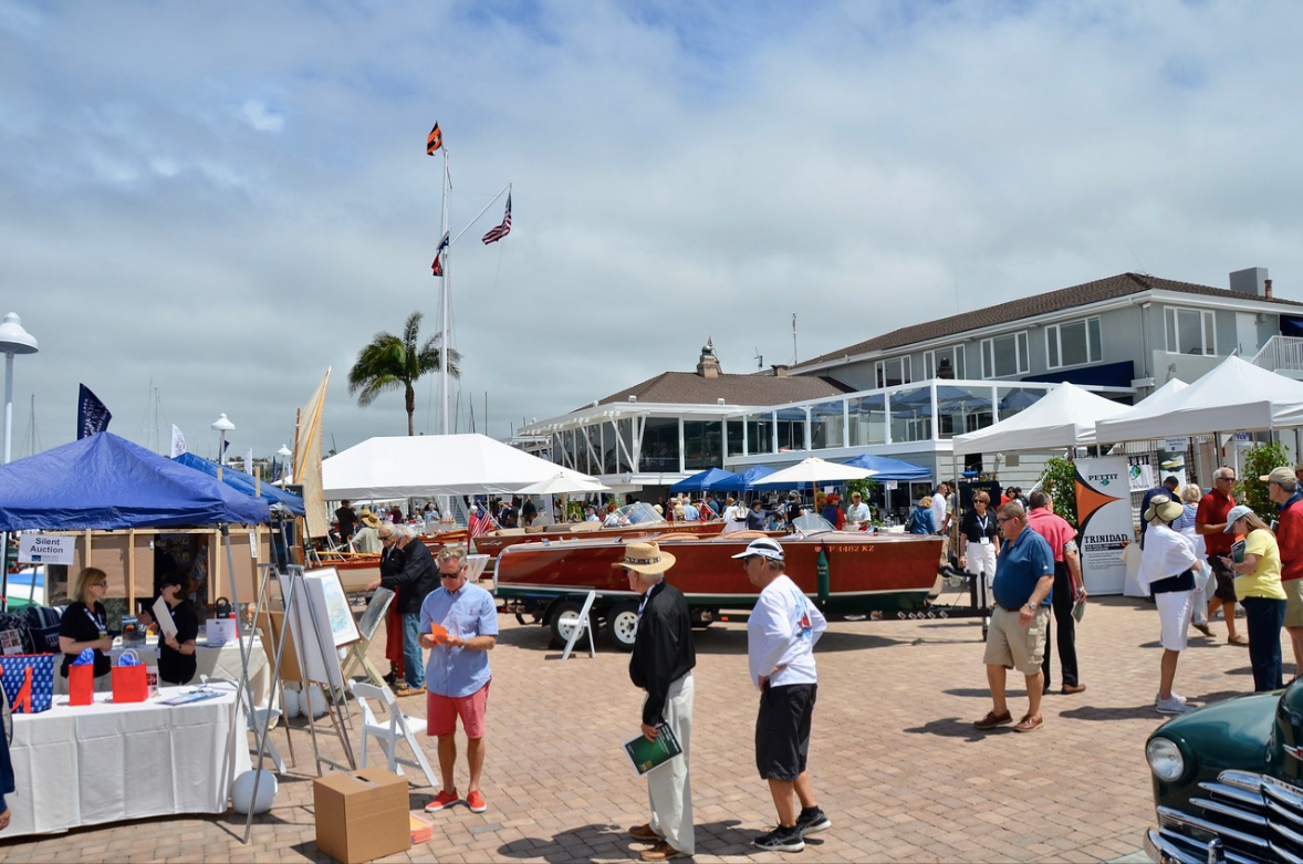 Outdoor market by the harbor with white tents, people browsing, and boats under a sunny sky