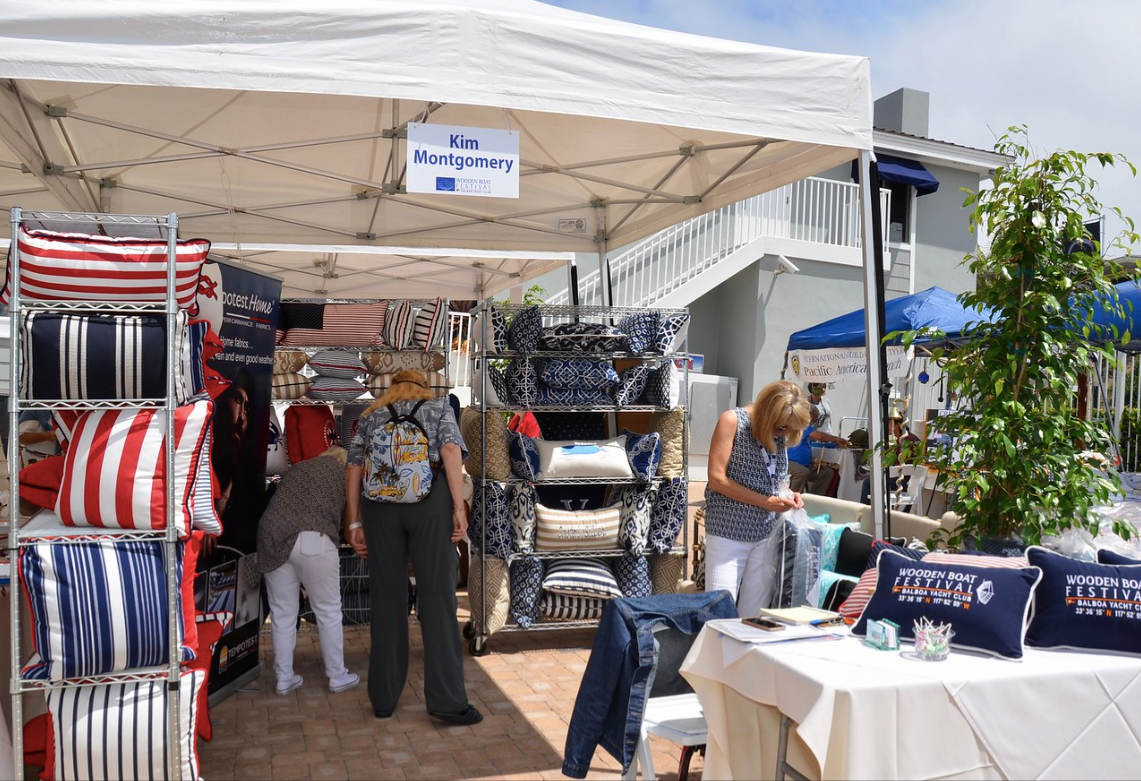 Outdoor market stall under a white canopy with hanging striped bags, clothes, and tables of merchandise