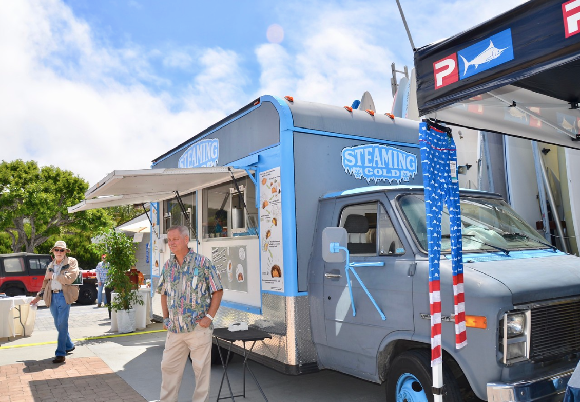Blue food truck at an outdoor event with people standing nearby and a market tent overhead