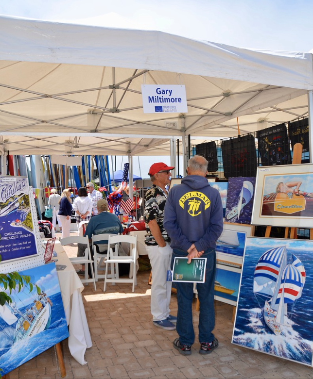 People browsing colorful art displays under a white canopy at an outdoor market booth.