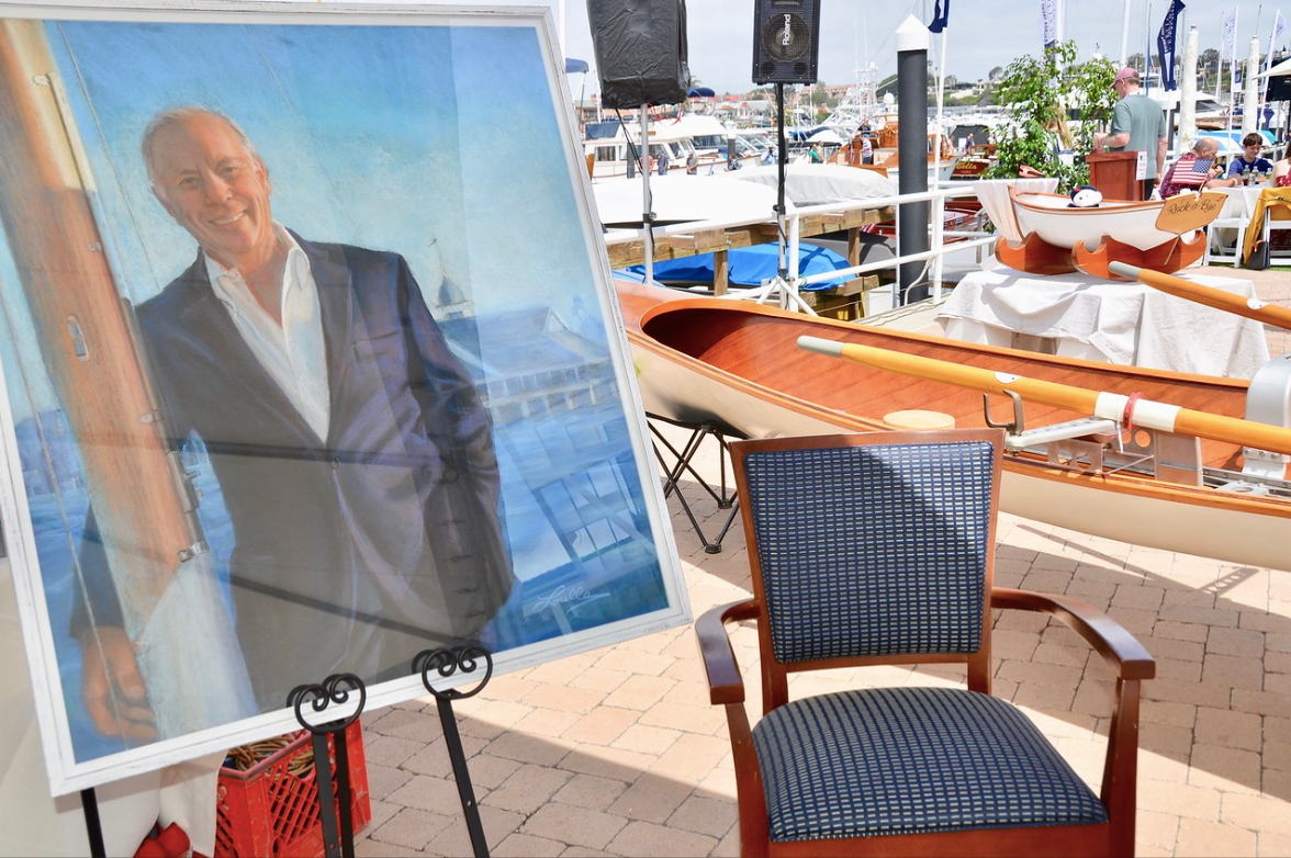 Framed portrait on an easel beside a striped chair at a sunny waterfront café terrace