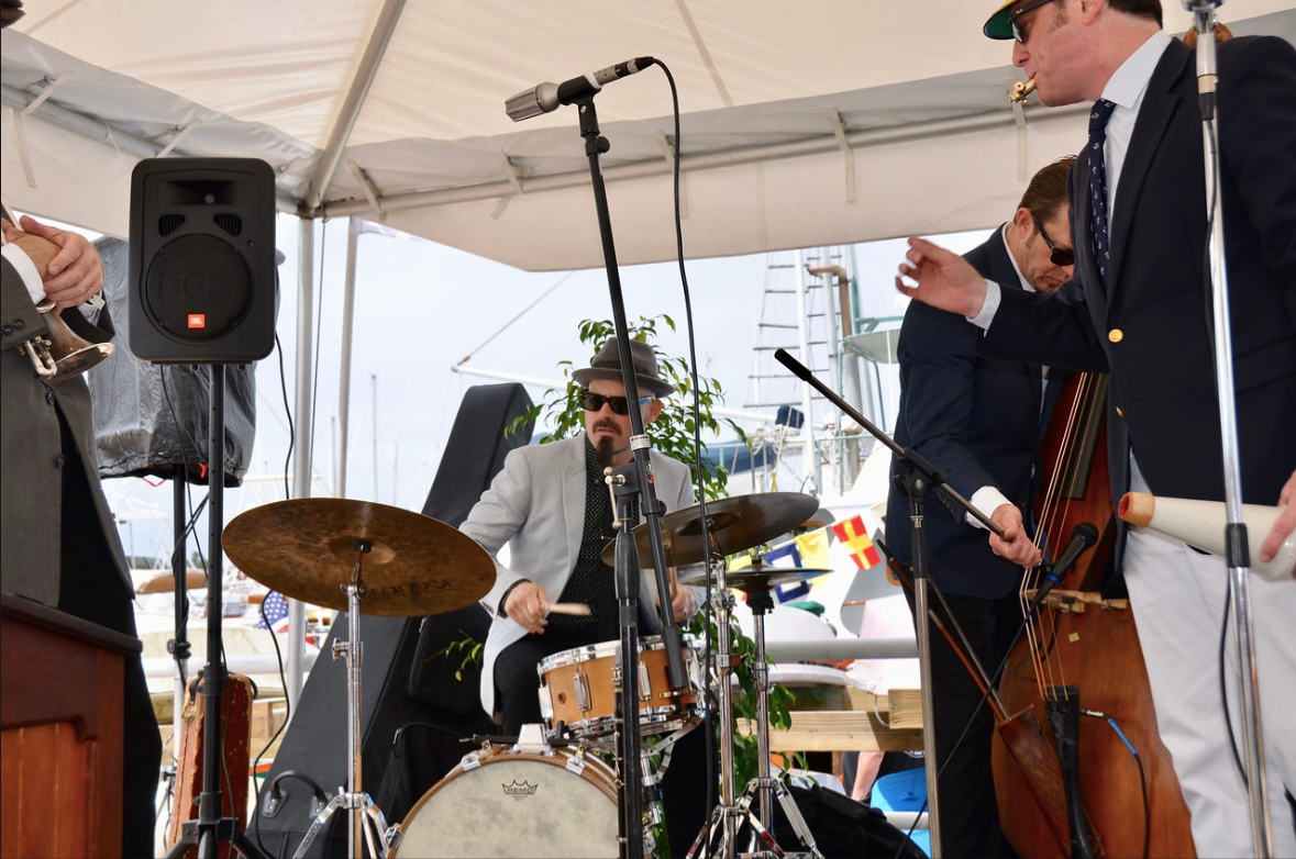 Live jazz band performing under a white tent with drums, bass, and microphones.