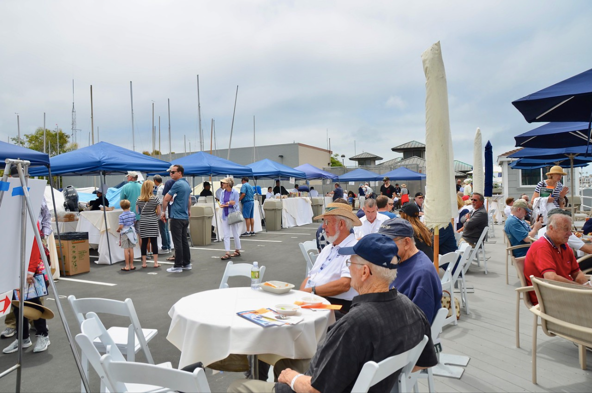 Outdoor marina café with people seated at white tables under blue umbrellas near the water