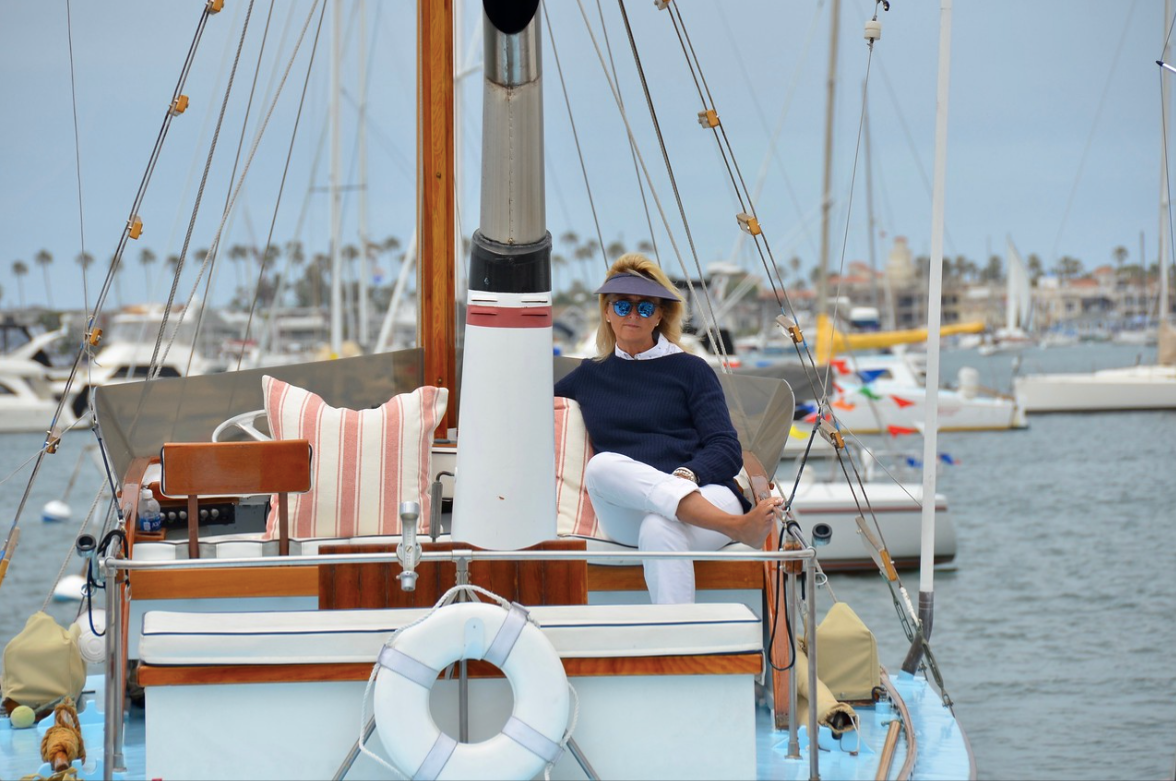 Person seated on a sailboat deck at a marina, with white life ring and masts in the background.