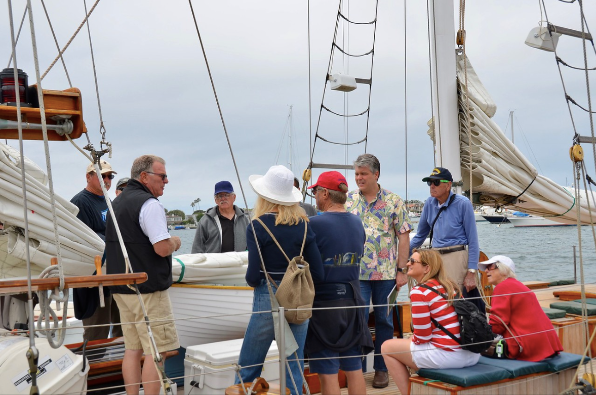 People gathered on a sailboat deck with ropes and masts, talking near the water.