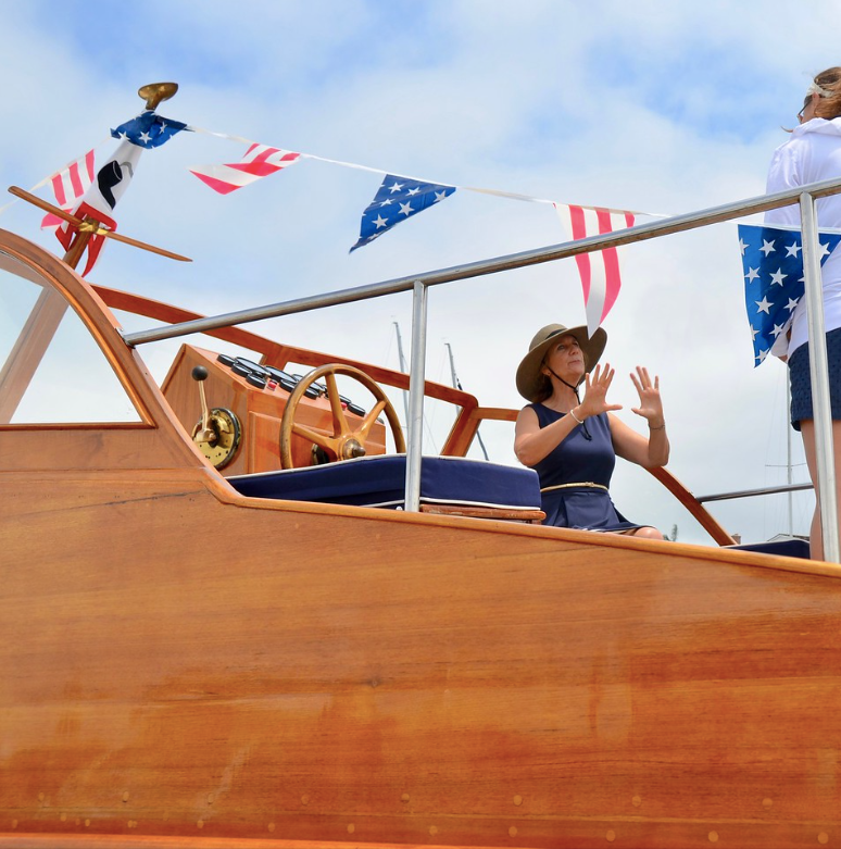 Person waving from a wooden boat decorated with red, white, and blue bunting under a blue sky