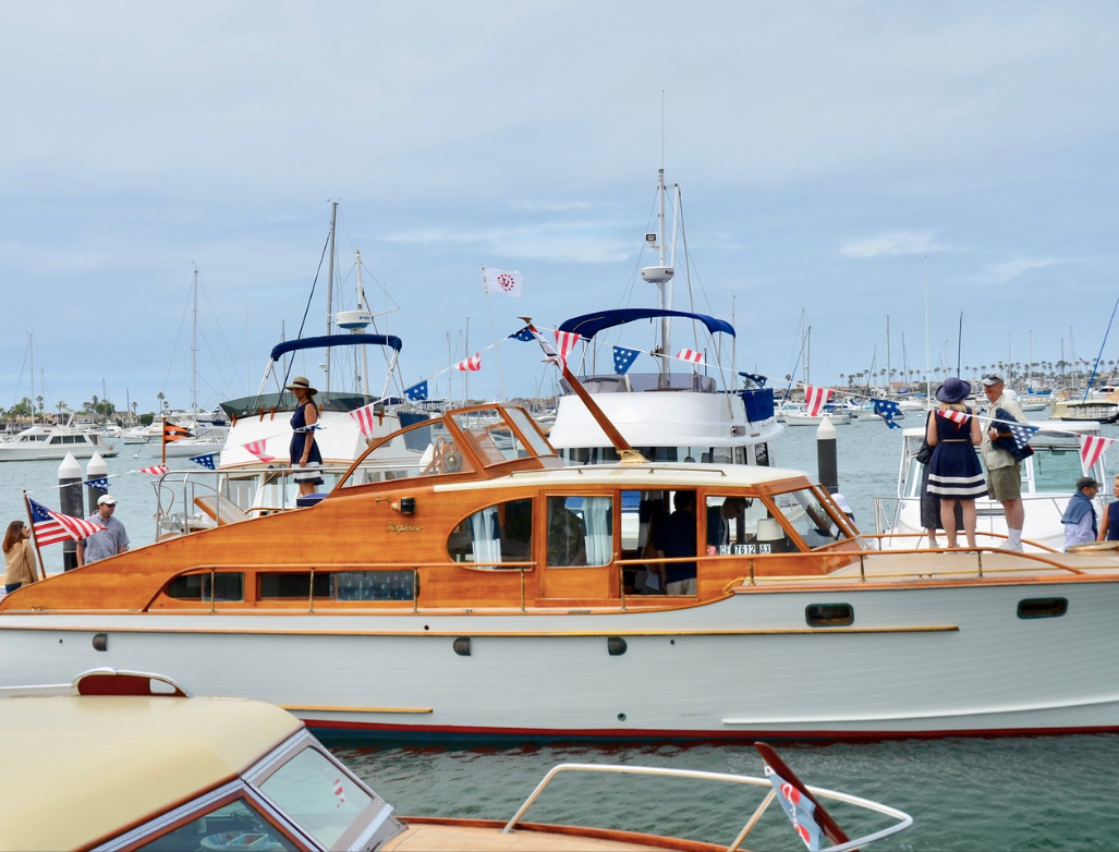 Wooden motor yacht docked at a harbor, with people standing on the pier and sailboats in the background.