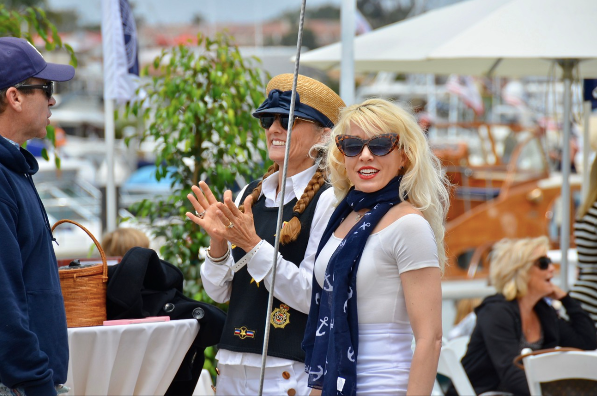 Two women in sunglasses smile at an outdoor market, one clapping beside a man under white umbrellas
