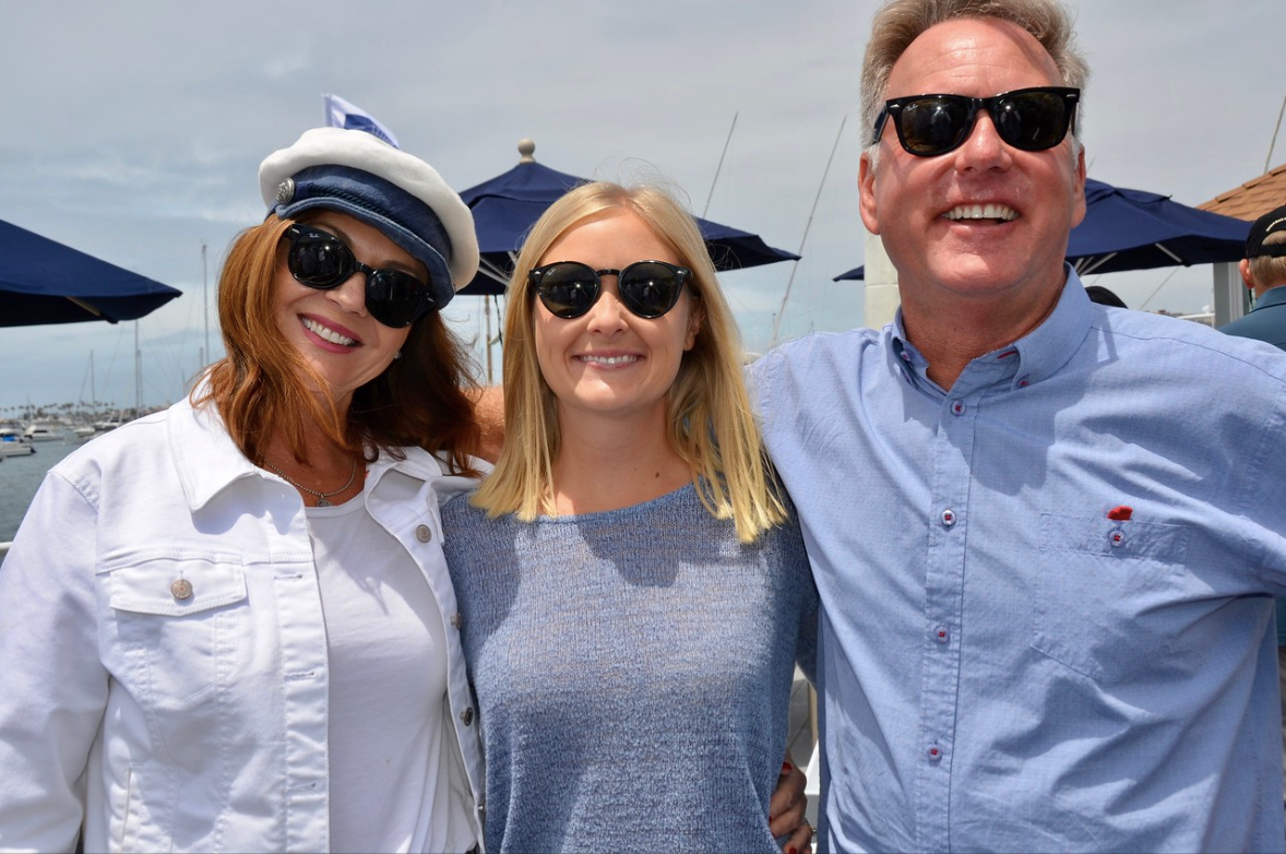 Three smiling people posing outdoors in sunglasses with blue umbrellas in the background