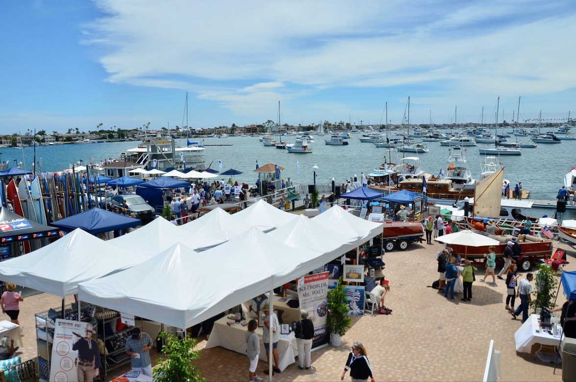 Crowded beach market with white tents, people, and many boats on calm blue water under a clear sky