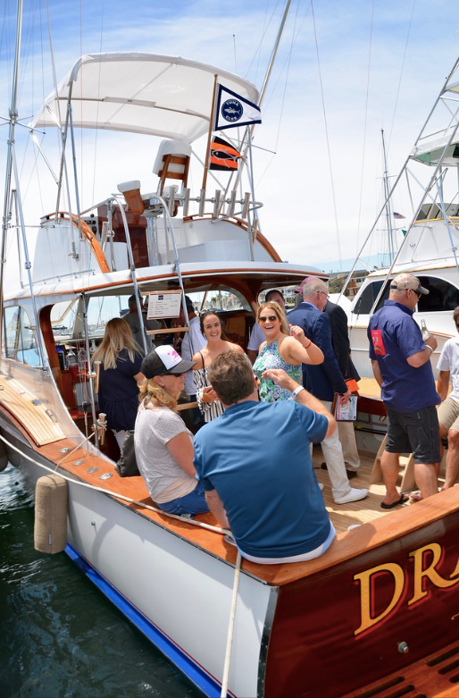 People gathered on a sailboat deck in a harbor, with white sails and blue water.