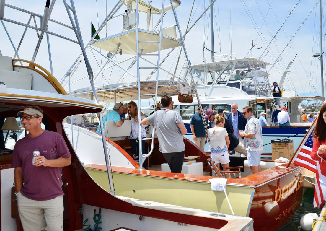 People gathering on boats at a busy marina, with masts, flags, and a bright blue sky