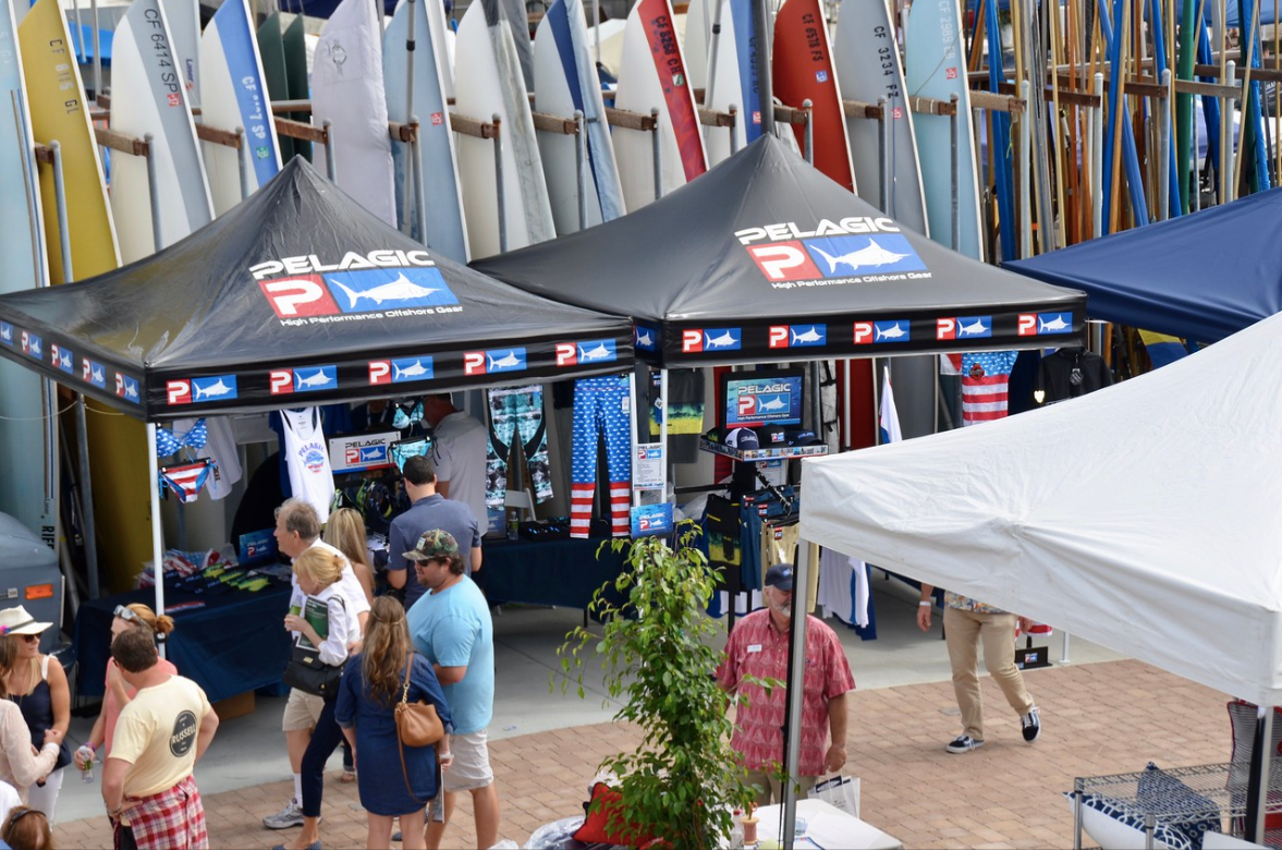 People under black tents at a busy outdoor event, with colorful sails in the background.