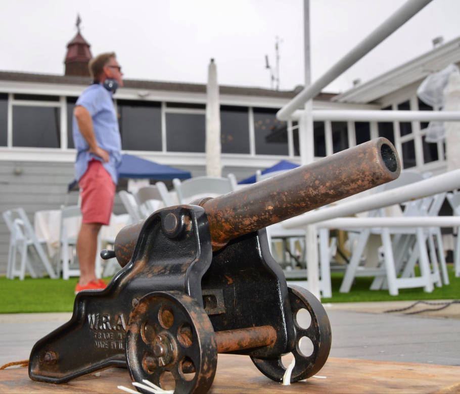 Rusty antique cannon mounted on a wooden table, with a person standing blurred in the background outdoors.