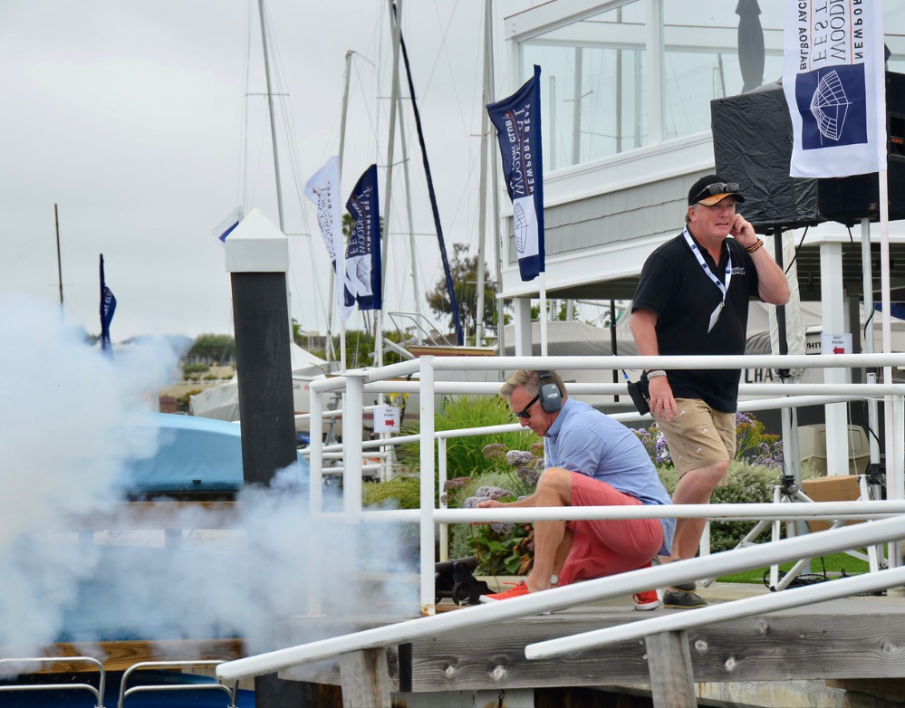 Two people on a dock near a smoking cannon, with blue flags and a boat in the background