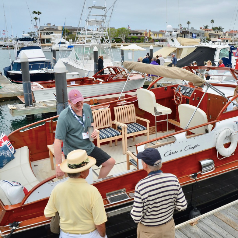 People chatting beside a red-and-white boat at a marina, with yachts and docks in the background