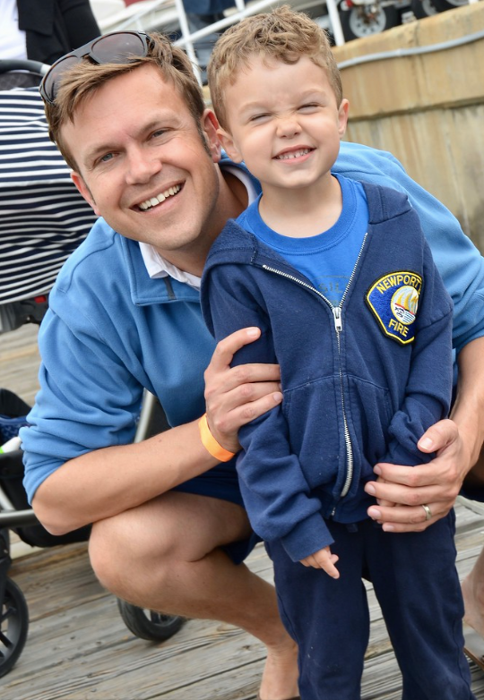 Smiling man crouches beside a child in a blue jacket outdoors on a wooden boardwalk